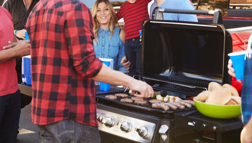 people standing around a barbecue grill that has sausages and burger patties. a green bowl of burger buns is next to the grill
